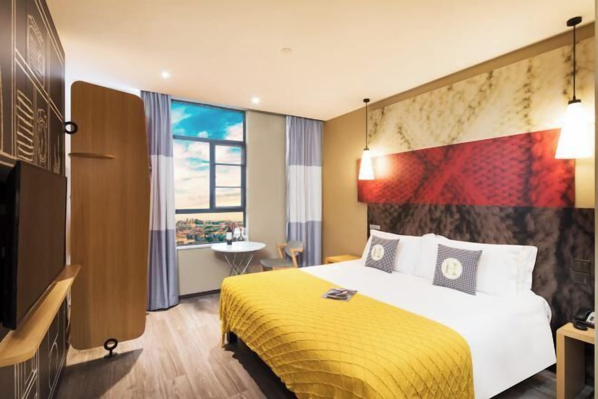 Una habitación de hotel con cama, ventana con vista a la ciudad, una mesita pequeña y una silla.