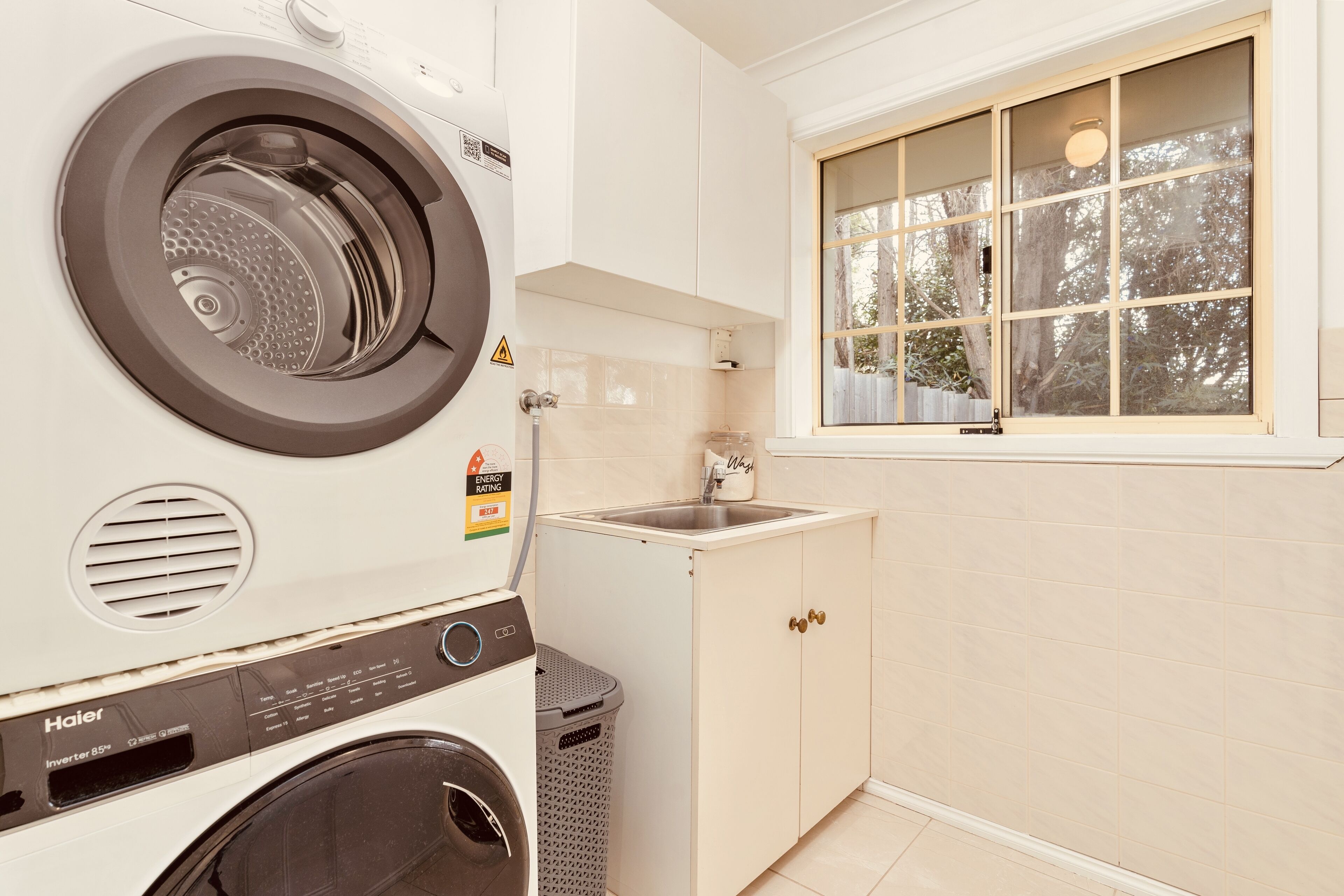 A laundry room with a front-loading washer and dryer, a sink, and a window with a view of trees.