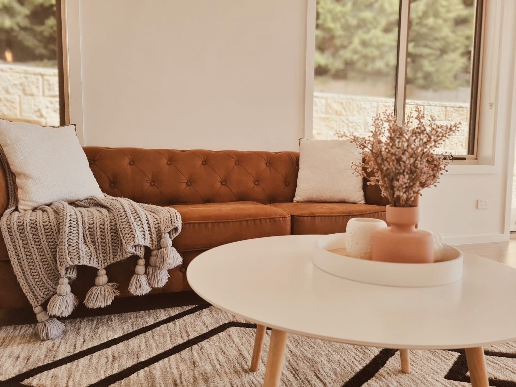 A living room with a brown sofa, white cushions, a knitted throw, a white coffee table, and a vase with dried flowers.