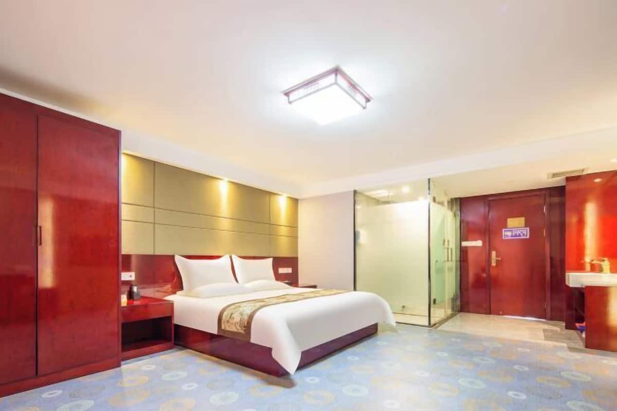 A hotel room with a large bed, a wardrobe, a bedside table, and a bathroom visible through a glass door.