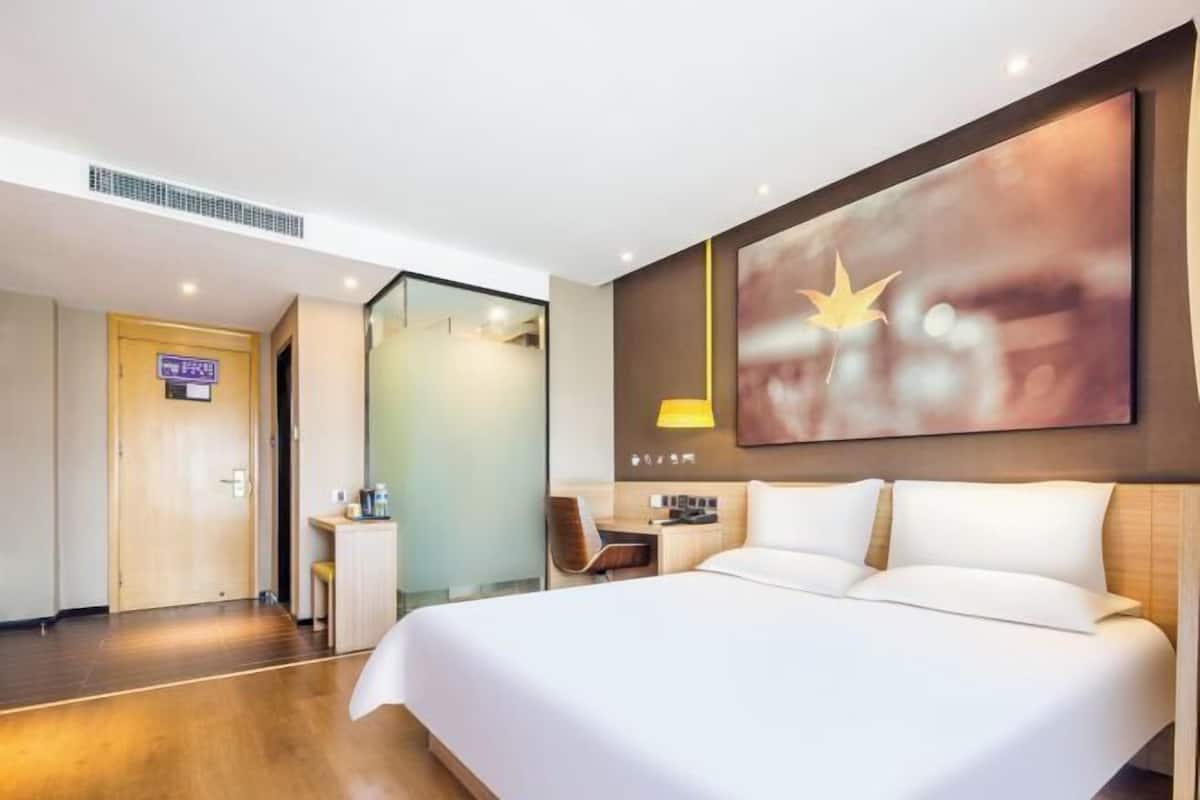 A hotel room with a large bed, a desk, a chair, and a wall-mounted lamp.