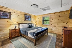 Quarto com painéis de madeira, uma cama, mesas de cabeceira, cômoda e televisão na parede.