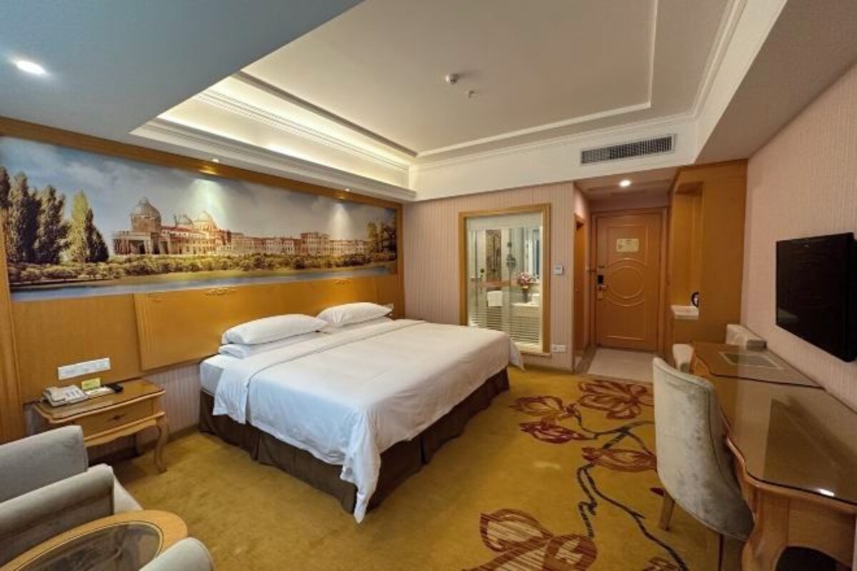 A hotel room with a large bed, a desk, a chair, a TV, and a mural on the wall.