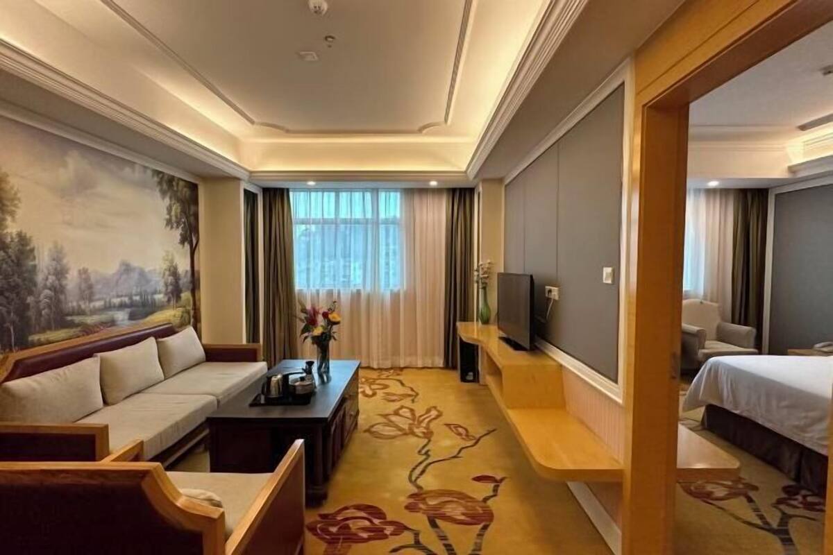 A hotel room with a sofa, a TV, a bed, and a painting on the wall.