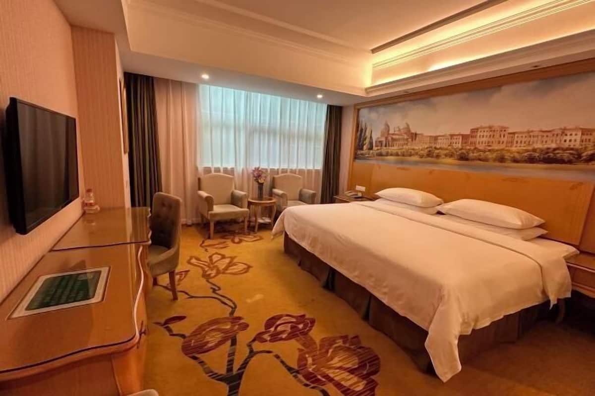 A hotel room with a bed, a desk, a TV, and a painting of a cityscape on the wall.