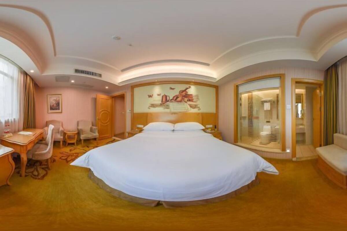 A hotel room with a large bed, a desk with a chair, a sofa, and a glass partition.
