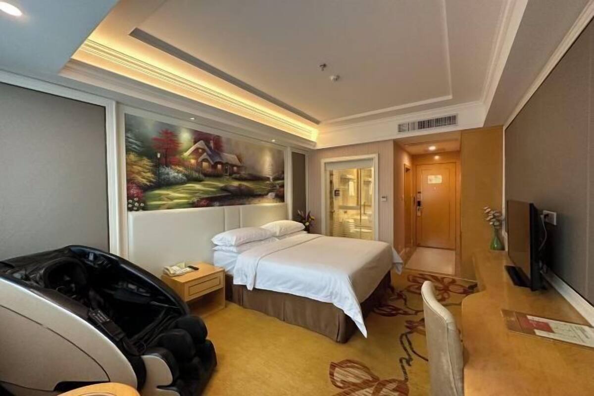 A hotel room with a bed, a massage chair, a painting on the wall, and a television.