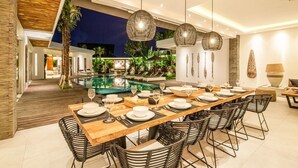 Ein moderner Essbereich mit einem langen Holztisch, gedeckt für eine Mahlzeit, umgeben von schwarzen Stuhlgestellen aus Draht, und einem Pool im Hintergrund.