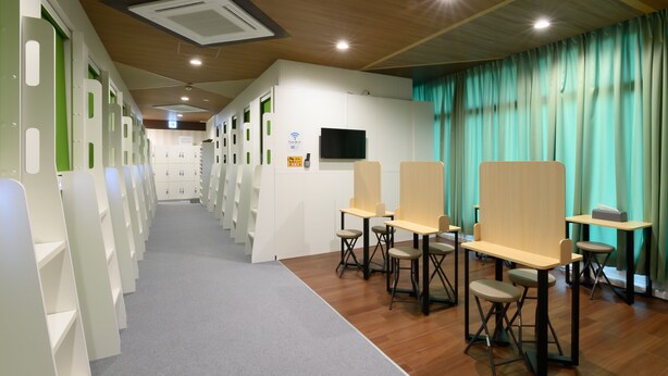 A modern, well-lit room with a long corridor, individual workstations with chairs, a flat-screen TV, and a sign indicating free Wi-Fi. - Tabist Capsule Inn Kasakake (Midori)