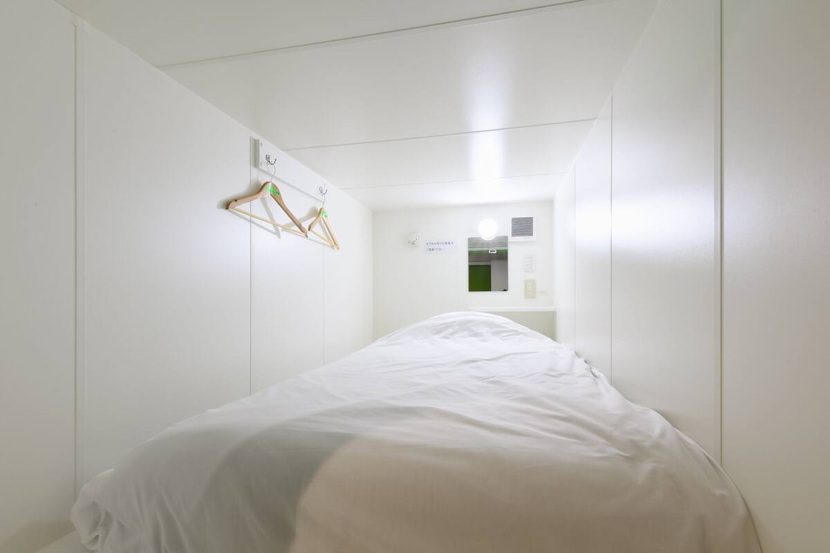 A minimalist hotel room with a single bed, a wall-mounted light, and a small mirror.