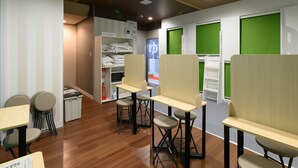A modern office space with wooden flooring, white walls, and green partitions. There are desks and chairs arranged for work, and a storage unit with folded clothes. - Tabist Capsule Inn Kasakake (Midori)
