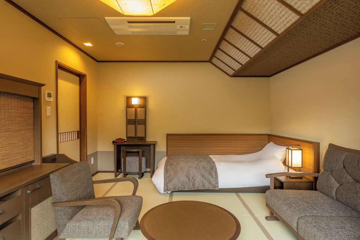 Ein Hotelzimmer mit einem Bett, zwei Stühlen, einem kleinen Tisch, einer Nachttischlampe und einem an der Wand montierten Heizkörper.