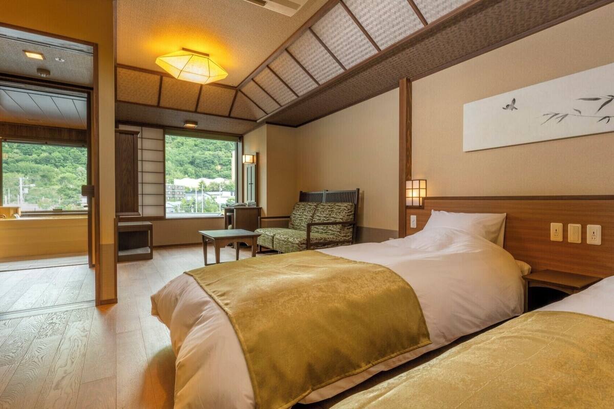 Ein Hotelzimmer mit zwei Betten, einer Sitzecke mit einem Sessel, einem großen Fenster und einem an der Wand befestigten Kunstwerk.