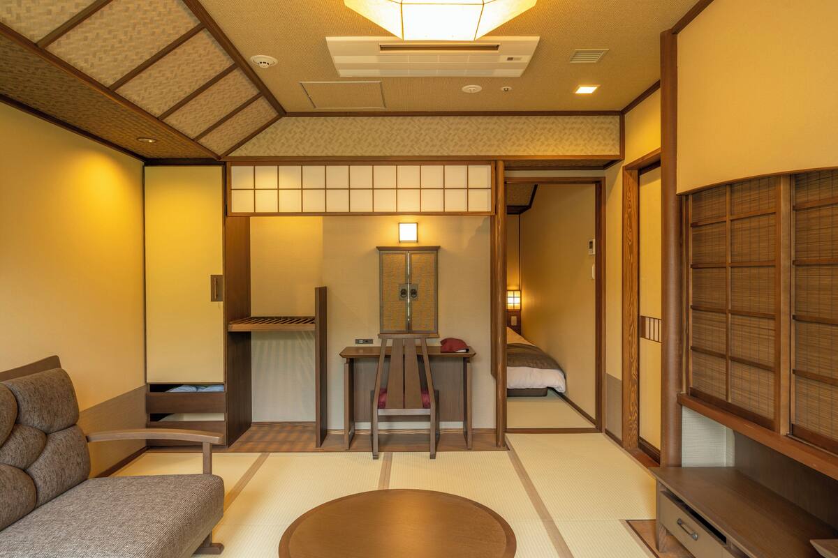 Ein traditioneller japanischer Raum mit Tatami-Boden, einem niedrigen Holztisch, einer gepolsterten Sitzgelegenheit und einer Schiebetür zum Schlafzimmer.