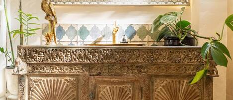 An ornate wooden cabinet with intricate carvings, a large mirror above it, and a potted plant on the right.