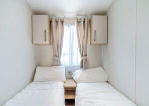 Quarto pequeno com uma cama de solteiro, um banco de madeira no centro, dois armários e uma janela com cortinas.