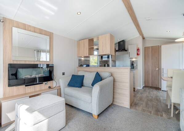 A modern living room with a sofa, ottoman, flat-screen TV, and a kitchen area with wooden cabinets and a microwave.