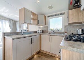 A modern kitchen with wooden cabinets, a built-in stove, a window with a view, and a countertop with a kettle. - Platinum Caravan 6 Berth (3 bed) (Pet) (Skegness)