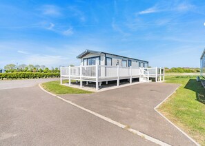 A single-story building with a covered porch and a small deck, surrounded by a paved driveway and grassy areas. - Platinum Caravan 6 Berth (3 bed) (Pet) (Skegness)