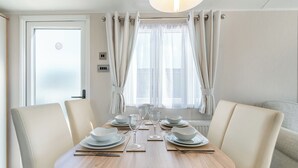 A dining area with a wooden table set for four, white chairs, and a window with curtains. - Platinum Caravan 6 Berth (3 bed) (Pet) (Skegness)