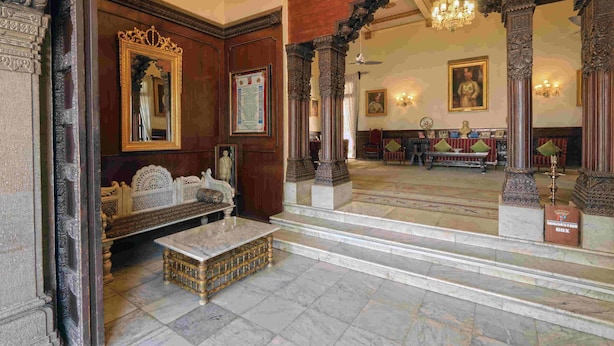 A grand lobby with ornate wooden columns, a marble floor, and a large mirror reflecting the room. - Nilambag palace (Bhavnagar)