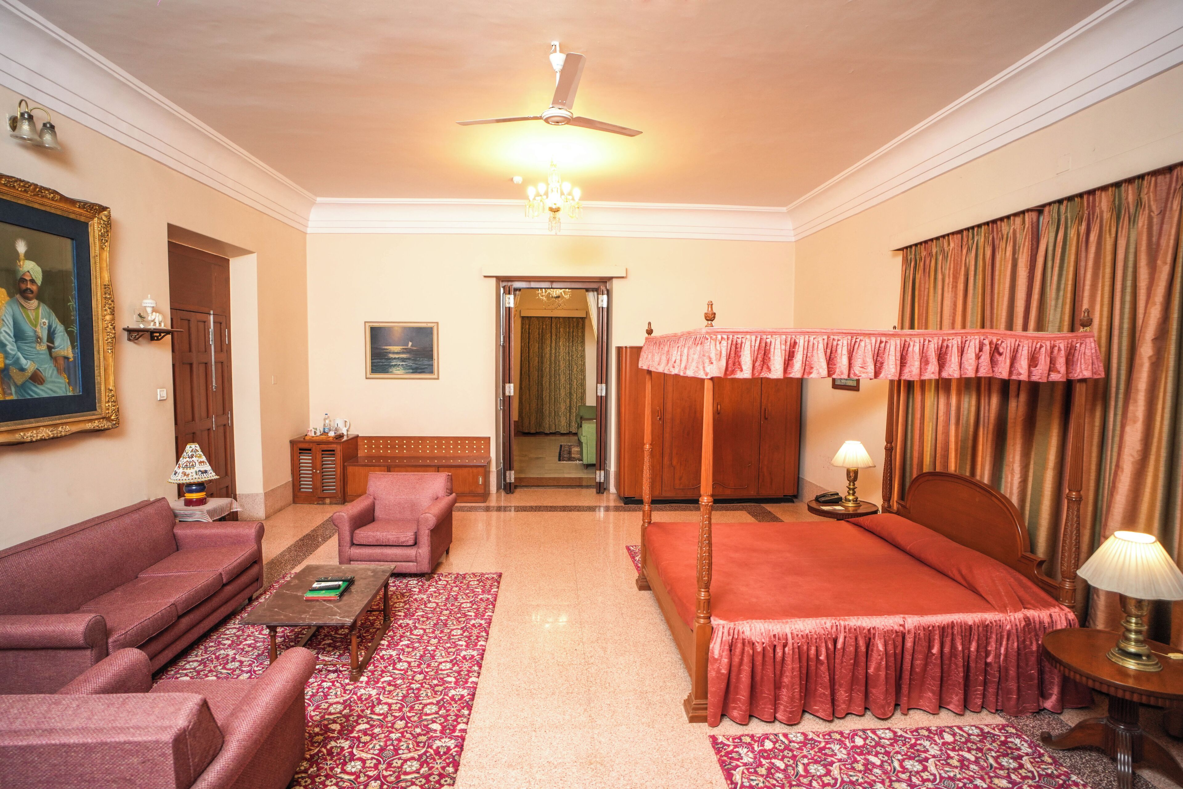 A hotel room with a four-poster bed, a sitting area with a sofa and armchair, a wooden cabinet, and a chandelier.