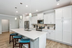 A modern kitchen with white cabinets, a large island with stools, stainless steel appliances, and pendant lighting.