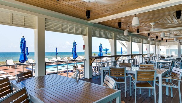 Un espace extérieur abrité, aménagé pour les repas, avec des tables et des chaises en bois, donnant sur une plage parsemée de parasols.