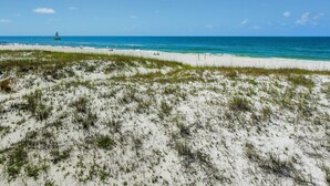 Une plage de sable blanc avec une végétation clairsemée, un ciel bleu azur et un voilier à l’horizon.