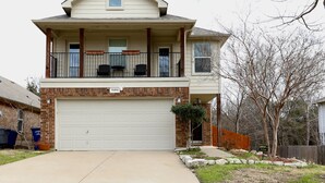 Uma casa de dois andares com garagem, varanda com móveis de exterior e uma pequena árvore no jardim da frente.