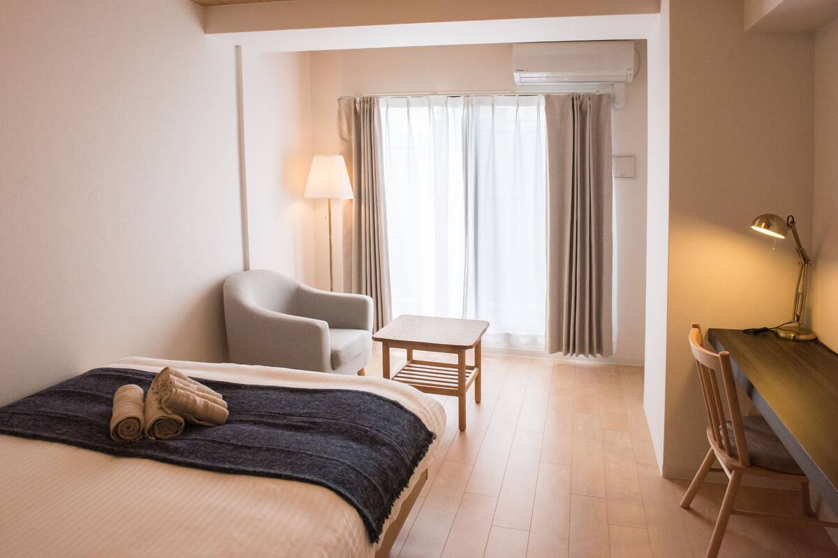 A hotel room with a bed, a chair, a small table, a desk, and a lamp.