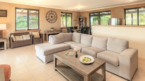 A modern living room with a sectional sofa, a wooden coffee table, and a dining area with a table and chairs. - Jamaicaville - 90 Acre hilltop off-grid eco-home with awesome views (Upper Growee)