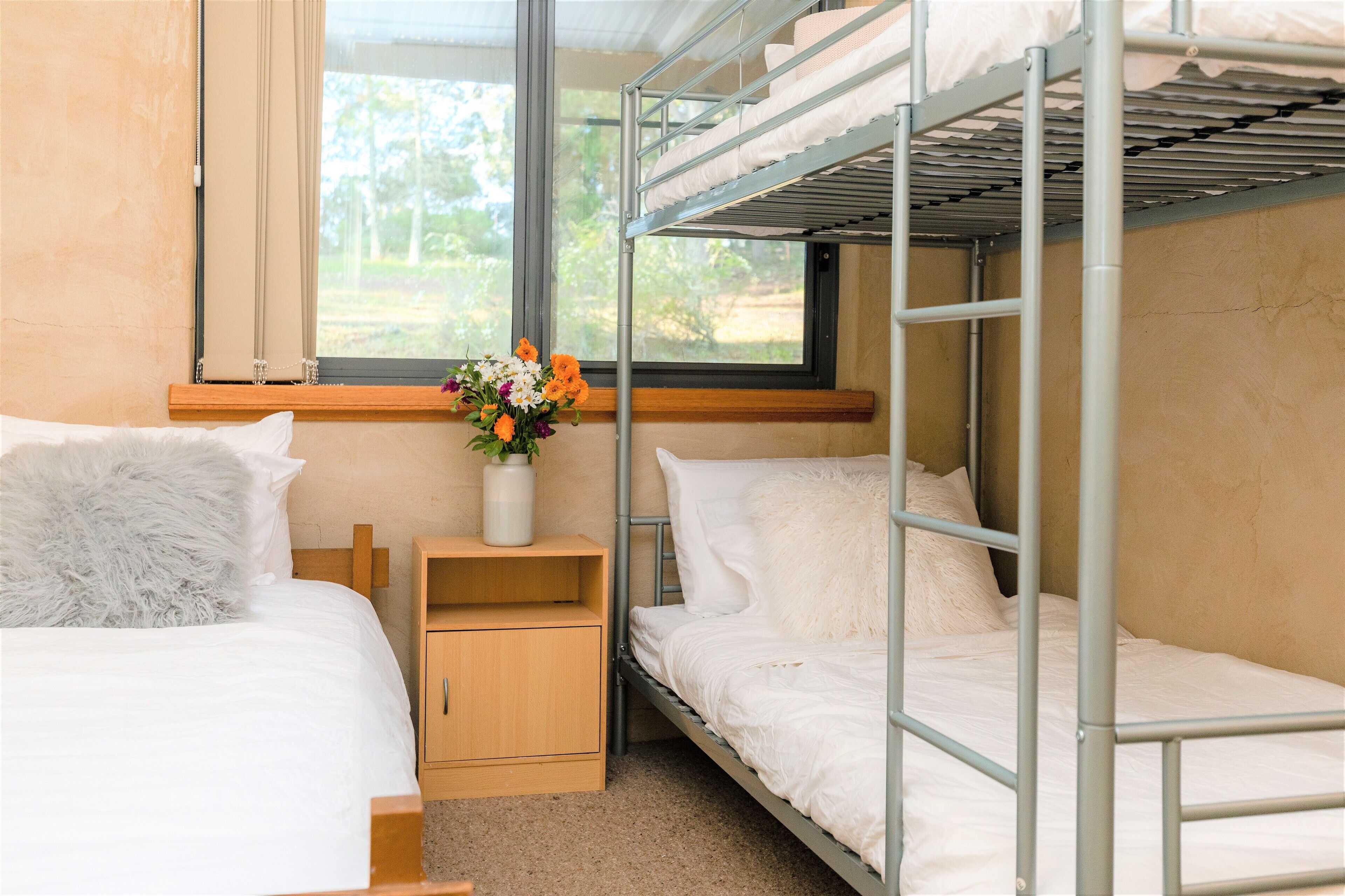 A bunk bed room with a window, a bedside table with a vase of flowers, and a wooden nightstand.