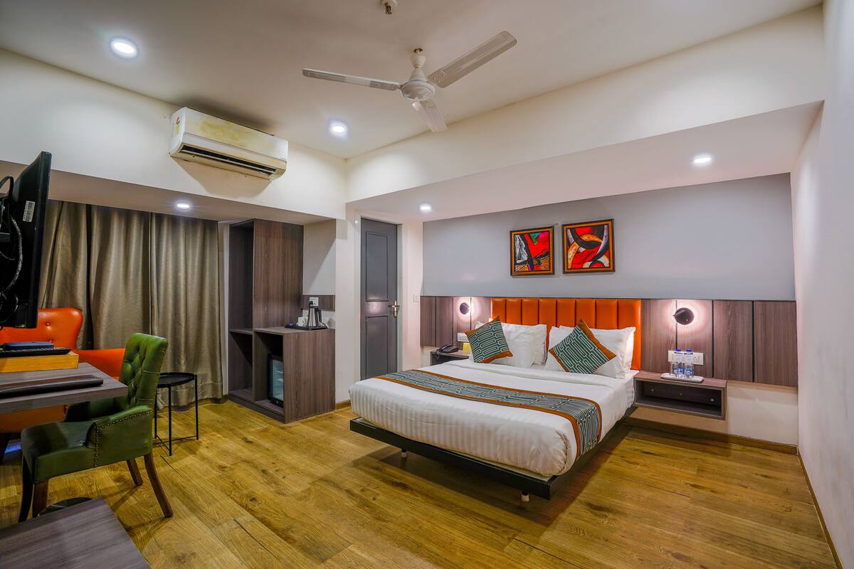 A modern hotel room with a bed, a desk, a chair, an air conditioner, a ceiling fan, and artwork on the wall.
