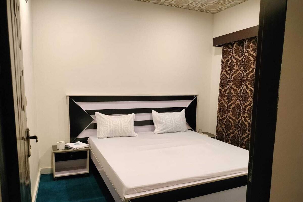 A neatly made bed with white linens and two pillows, a bedside table with a lamp, and a patterned curtain on the window.