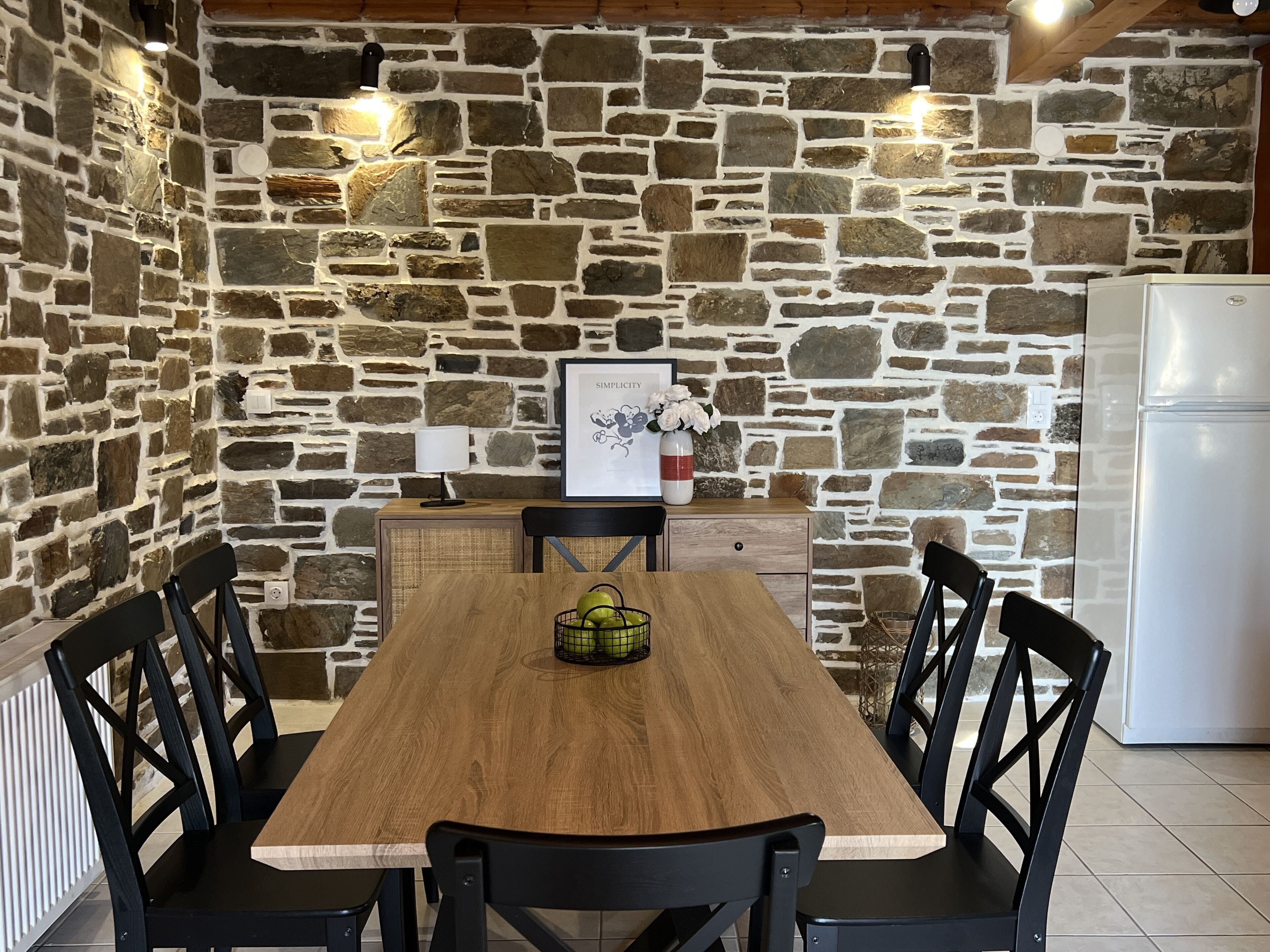 Un coin repas avec une table en bois et des chaises noires, un mur en pierre, un réfrigérateur blanc et un tableau encadré accroché au mur.