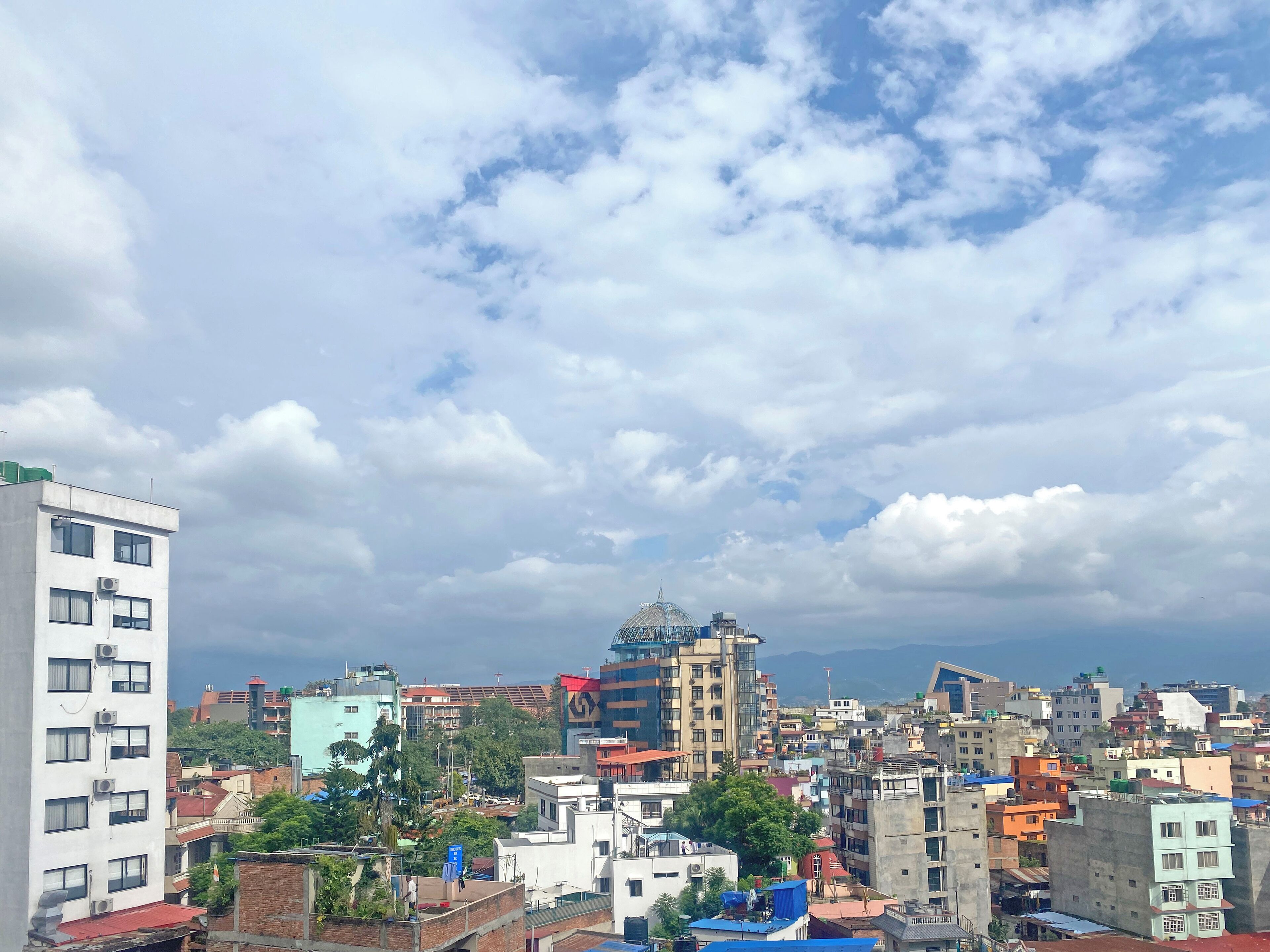 A cityscape with various buildings, including a prominent white building with multiple air conditioning units and a blue-domed structure.