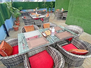 An outdoor seating area with wicker chairs and red cushions, surrounded by potted plants and a green wall.