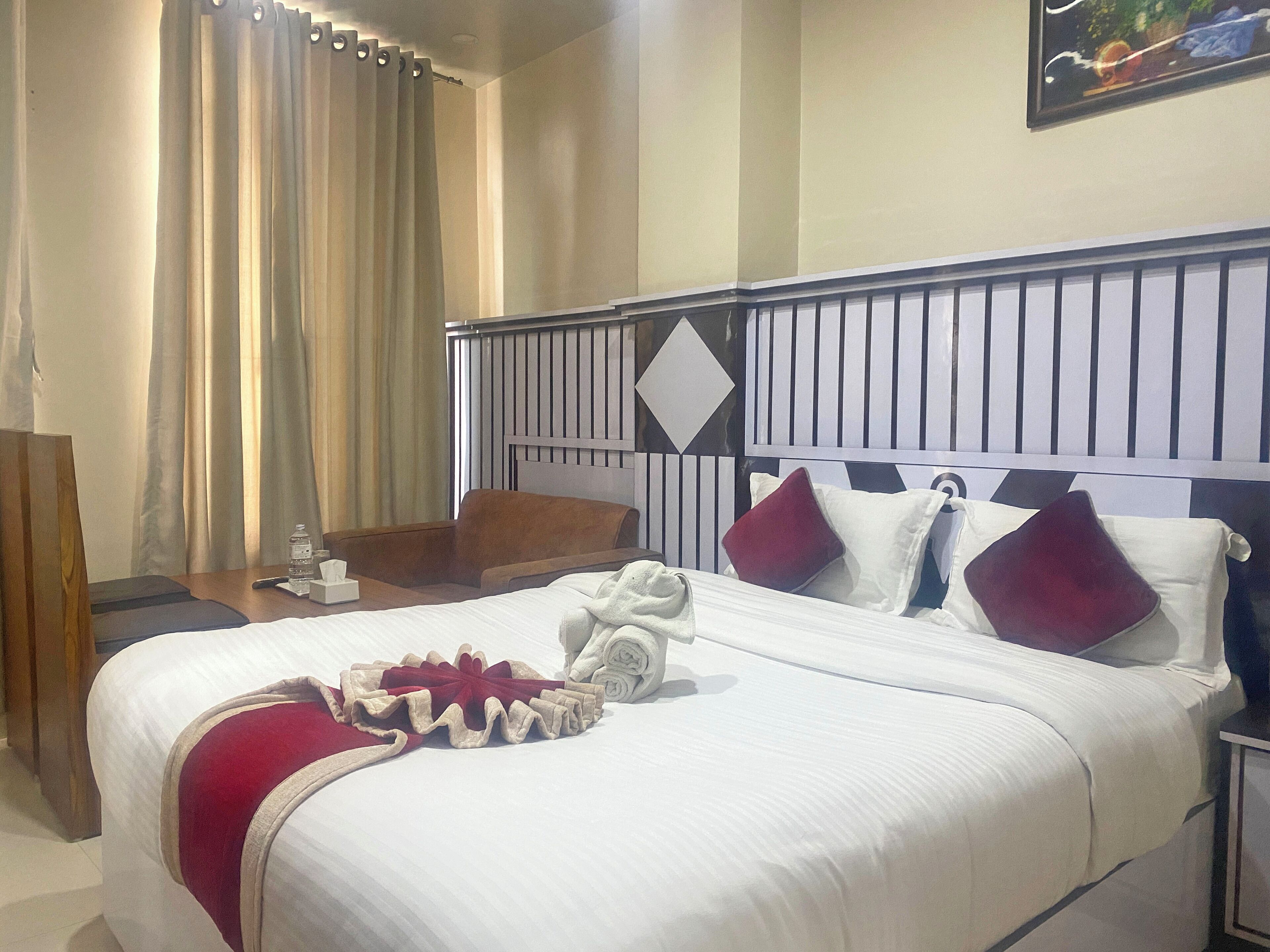 A hotel room with a neatly made bed, a wooden side table, a water bottle, and a painting on the wall.