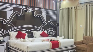 Una habitación de hotel moderna con una cama grande, dos sillones, una mesita con una botella de agua y un cabecero decorativo.
