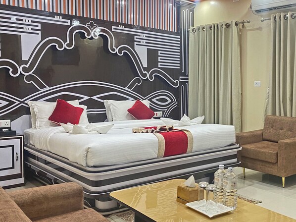 A modern hotel room with a large bed, two side chairs, a small table with a bottle of water, and a decorative headboard.