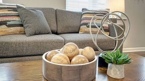 Un tazón de madera con bolas de madera sobre una mesa de madera, un sofá gris con cojines a rayas y una pequeña planta en maceta en la esquina.