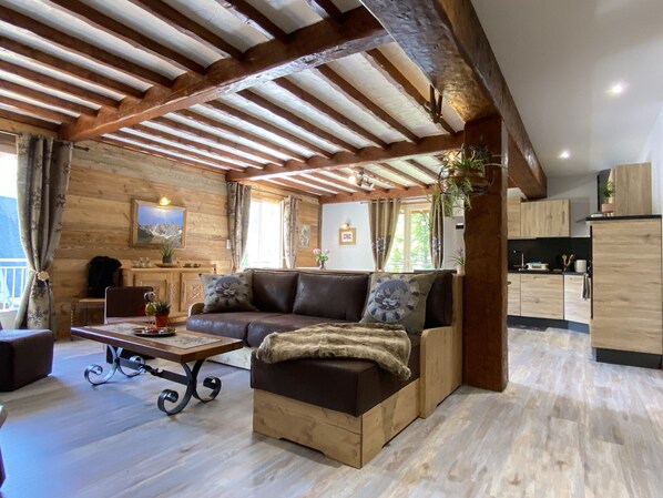 A cozy living room with a sofa, a coffee table, and a dining table. There is a kitchen area with wooden cabinets and a refrigerator.