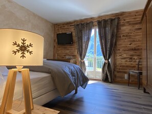 A bedroom with a wooden wall, a bed, a TV mounted on the wall, and a window with curtains. - Vaste T4 115m2. Balcon. Vue. Suite. Jardin et jeux (Cauterets)