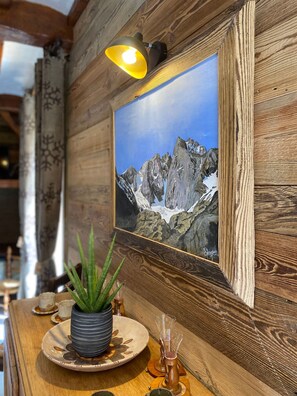 A wooden wall with a framed picture of a mountain landscape, a potted plant, and a table with a plate and glasses.
