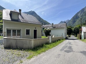 Maalaismaisemassa kulkeva tie johtaa pieneen, kaltevakattoiseen rakennukseen, jonka ympärillä kohoavat vuoret.
