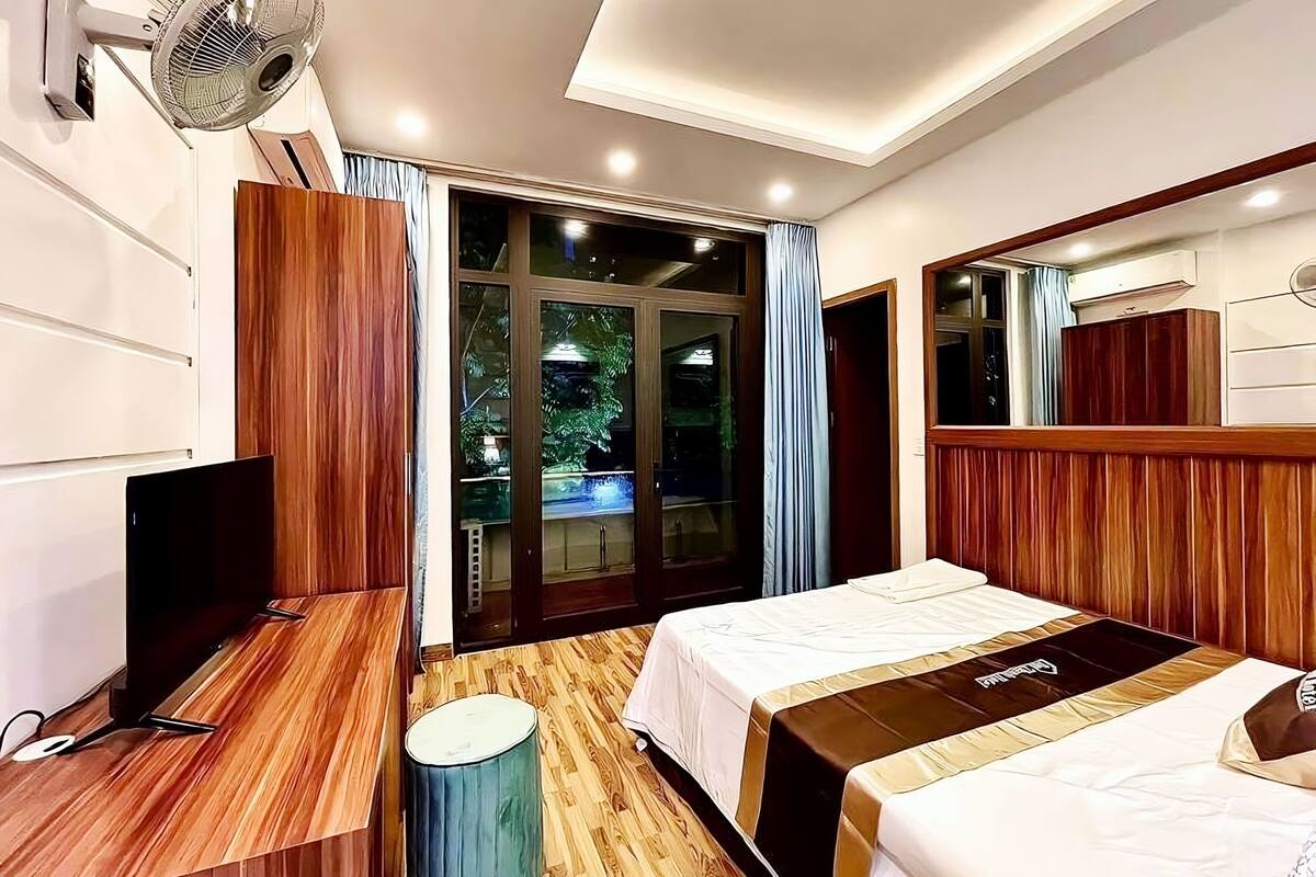 Ein modernes Hotelzimmer mit Holzboden, einem Bett mit gestreifter Bettdecke, einem Schreibtisch mit Computer, einem Standventilator und einem großen Fenster mit Vorhängen.