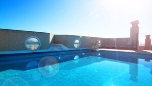 Una piscina azul cristalina con ventanales circulares en el borde, un cielo iluminado por el sol y un edificio lejano.