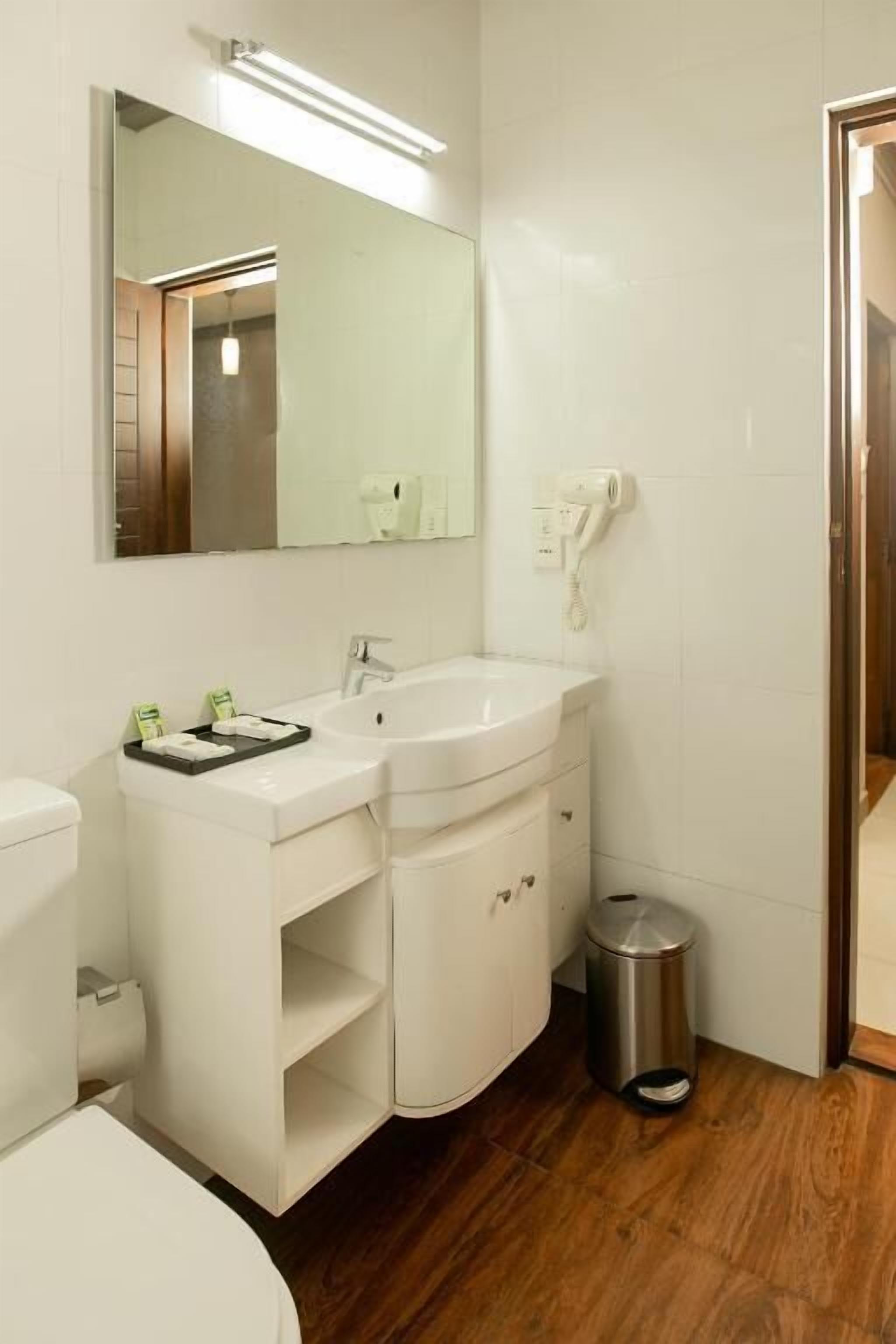 Un baño con lavamanos blanco, espejo e inodoro. Hay un cubo de basura y un secador de pelo en la pared.