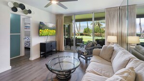 Ein modernes Wohnzimmer mit großem Fernseher, einem Glastisch, einer Couch und einem Deckenventilator.